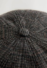 Grå ullflat cap med ett texturerat mönster, har en centrerad knapp och sydda paneler. Tygdet verkar mjukt och något fluffigt.