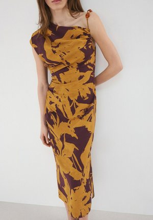 Mujer que lleva un vestido midi sin mangas con un estampado floral abstracto en color amarillo mostaza y púrpura oscuro, con un escote asimétrico y tirante en el hombro.