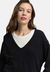 Vrouw met lang bruin haar die een wit crewneck shirt draagt, laag onder een zwarte V-halstrui tegen een effen achtergrond.