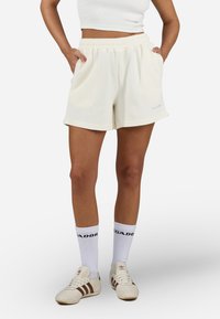 Shorts boje kreme s rebrastim uzorkom, elastičnim pojasom, bočnim džepovima i malim logom na rubu. Upareni s bijelim čarapama i cipelama.