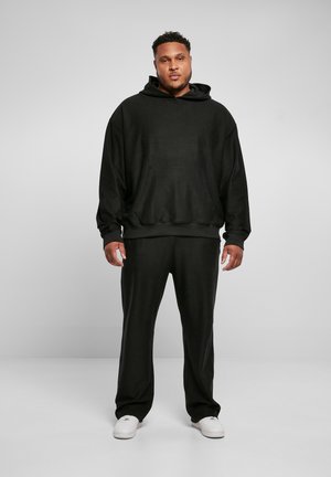 Haut de survêtement noir texturé avec une poche avant et des poignets côtelés, associé à un pantalon noir assorti. Des baskets blanches complètent la tenue.