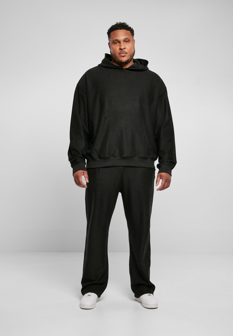 Homme debout portant un sweat à capuche noir et un pantalon noir assorti avec des baskets blanches sur un fond clair uni.