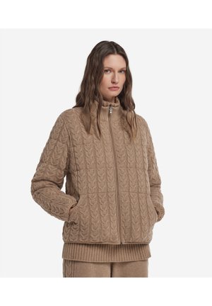 Vrouw draagt een bruine kabelgebreide jas met rits en hoge kraag, handen in de zakken en een bijpassende geribde trui eronder.