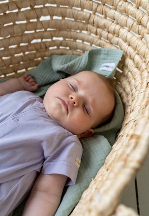 A light purple cotton outfit with short sleeves, resting on a green textured blanket inside a woven bassinet.
