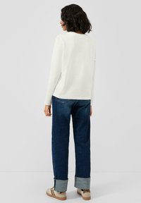 Biała koszula z długim rękawem, połączona z ciemnymi dżinsami z podwiniętymi mankietami i beżowymi tenisówkami, widziana od tyłu. Gładka faktura na całej powierzchni.