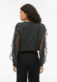 Blusa nera a pois con maniche trasparenti e arricciate, caratterizzata da un collo rotondo e una vestibilità rilassata. Abbinata a pantaloni neri a vita alta.