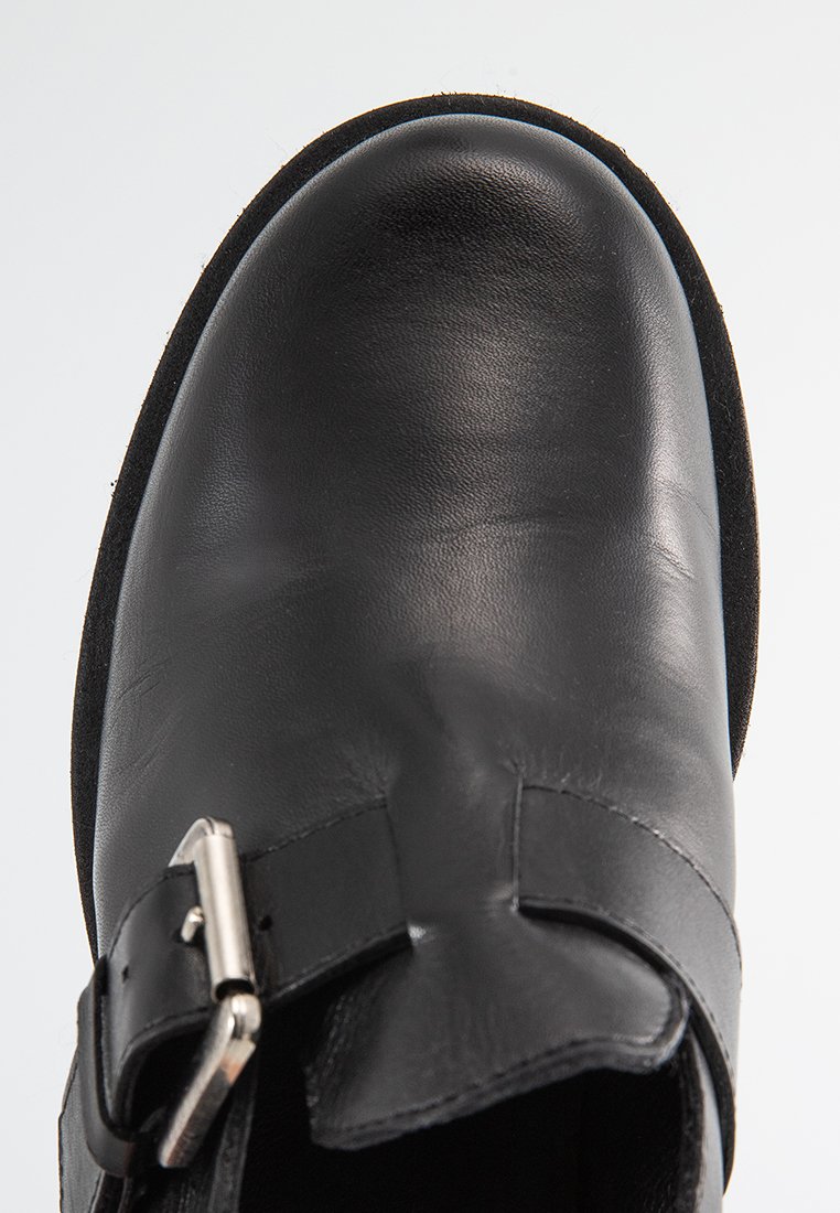 Chaussure en cuir noir avec un bout rond, surface lisse et une boucle en métal. La semelle est en matériau noir texturé.