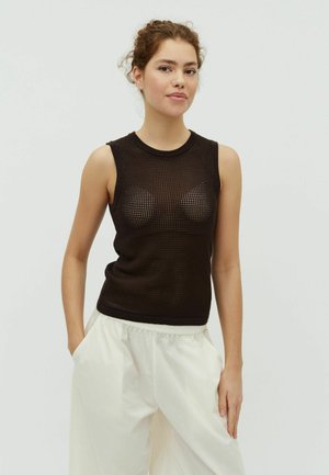 Jeune femme portant un haut en maille noire sans manches et un pantalon blanc ample, debout avec une main dans la poche devant un fond uni.