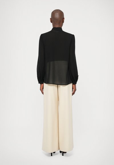 Melna caurspīdīga blūze ar puffadām piedurknēm un mežģīņu aprocēm, kombinēta ar plašām krēmkrāsas biksēm. Apģērbs izceļas ar minimālistisku dizainu un gludām tekstūrām.