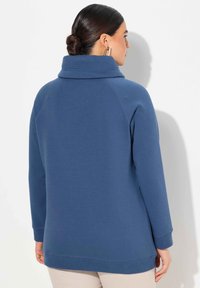 Felpa blu con colletto alto, maniche lunghe e polsini a coste. Presenta una vestibilità rilassata e una texture morbida, mostrata da dietro.