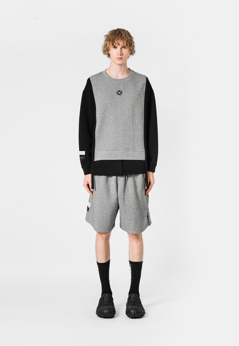 Sudadera gris con mangas negras, con un logo en el frente. Combinada con pantalones cortos grises, detalle de cadena, y calcetines negros con calzado negro.