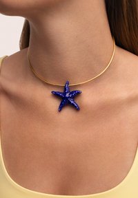 Le collier ras du cou en or présente un pendentif en forme d'étoile de mer bleue. L'étoile de mer est texturée avec une finition lisse et brillante, contrastant avec le collier minimaliste.