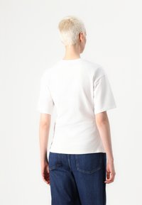 Tricou alb din bumbac, cu mâneci scurte, guler rotund și croială lejeră, văzut din spate, asortat cu pantaloni din denim albastru.
