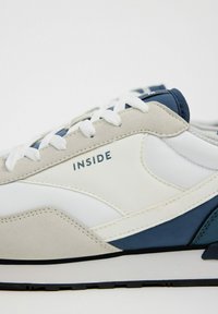 Zapatilla con una mezcla de cuero blanco, ante gris y acentos en azul marino. Presenta cordones planos blancos y la palabra "INSIDE" impresa en el costado.