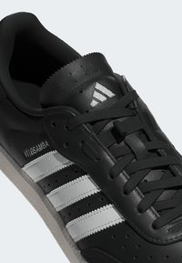 Czarne skórzane sneakersy z białymi paskami, teksturowanym językiem, okrągłymi sznurówkami i perforacjami po bokach. Posiadają branding "VELOSAMBA".