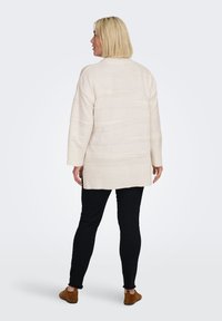 Pull crème long avec un motif texturé, poignets et ourlet côtelés. Associé à un pantalon noir et des chaussures en daim marron.