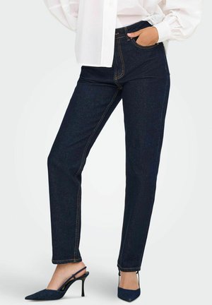 Jean en denim bleu foncé à taille haute, coupe droite et coutures jaunes visibles. Accompagné de talons noirs à talon ouvert.