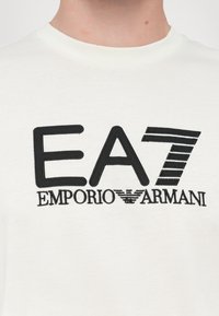 White cotton t-shirt featuring a large black embroidered "EA7" logo with "EMPORIO ARMANI" text below. Simple crew neckline.