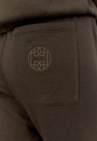 Tmavě hnědé tepláky s hladkou strukturou, mají boční kapsu a vyšívané kulaté logo ve světlejším odstínu.