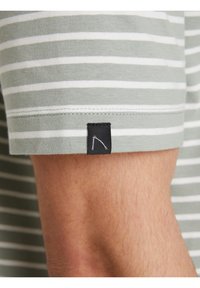 Camisa de manga corta a rayas con rayas horizontales en tonos verde apagado y blanco, con una etiqueta negra en el puño que muestra un sencillo logo en blanco.