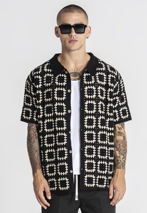 Mann mit kurzem Haar und Tattoos trägt große schwarze Sonnenbrille, ein kurzärmeliges Hemd mit schwarz-weißem Muster über einem weißen Tanktop.