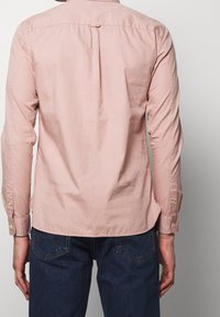 Camisa de manga larga rosa claro con canesú trasero y puños abotonados, combinada con jeans de mezclilla azul oscuro con bolsillos decorados con patrones.