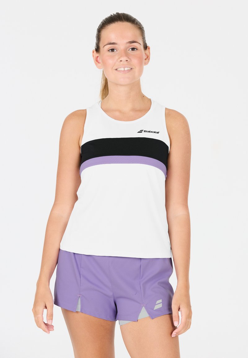 Giovane donna in top senza maniche bianco con strisce nere e viola e pantaloni sportivi viola, in piedi contro uno sfondo bianco.