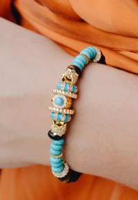 Bracelet de perles turquoise avec accents dorés, présentant des éléments décoratifs et des ornements en strass. Des perles noires et brunes ajoutent du contraste.