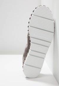 Chaussure en daim gris avec une surface texturée et des clous ronds le long du bord, dotée d'une semelle en caoutchouc blanc avec des rainures horizontales.