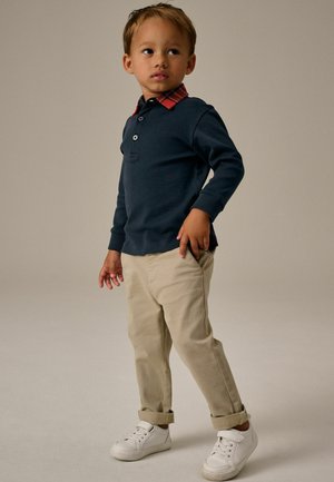 Polo a maniche lunghe blu navy con colletto a quadri rossi, abbinato a pantaloni beige e scarpe da tennis bianche. Tessuto morbido e design classico.