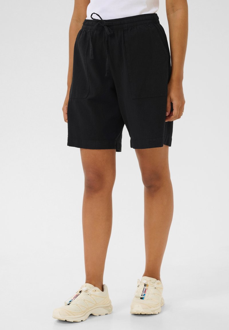 Shorts en coton noir avec taille à cordon, deux poches avant, design long jusqu'aux genoux et coupe décontractée. Associés à des baskets beiges.