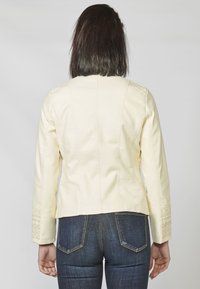 Giacca color crema con un design aderente, dettagli testurizzati sulle spalle e accenti ricamati sulle maniche. Indossata con jeans scuri.
