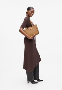 Sac fourre-tout en cuir marron au design structuré, avec des poignées doubles et un accent logo sur le devant. Porté avec une robe marron ajustée.