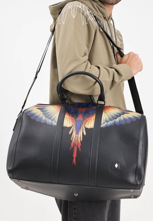 Uomo con felpa con cappuccio verde oliva che porta una borsa da viaggio in pelle nera con un design colorato di ali di uccello sulla parte anteriore e manici neri.