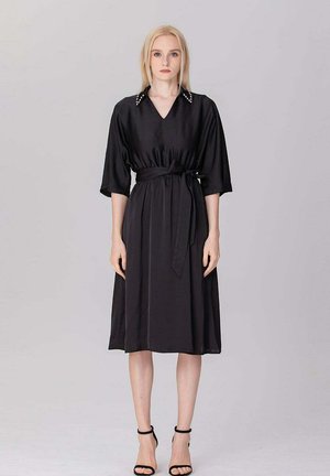 Femme blonde debout, portant une robe midi noire avec une taille ceinturée, des manches mi-longues, un col orné de perles et des talons noirs à bride cheville sur un fond uni.