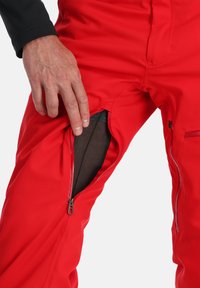 Rote Skihose mit einer seitlichen Reißverschlusstasche, sichtbarem Netzfutter und einem schlanken Design. Der Stoff wirkt glatt mit einer wasserabweisenden Beschichtung.