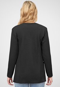 Langermet svart genser med teksturert overflate, buet hem og sidespalter. Vynes fra baksiden med rund hals.