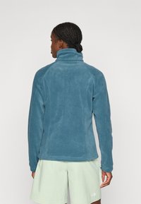 Veste en polaire teal avec un col haut et des manches longues. Présente une texture lisse et une coupe ajustée. Portée par-dessus un short vert clair.