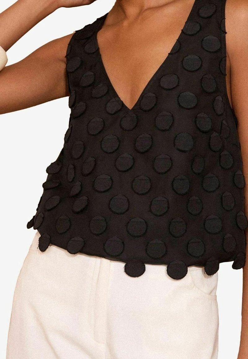 Haut noir sans manches avec motifs circulaires texturés porté avec un pantalon blanc, présentant un décolleté en V profond et une coupe décontractée.