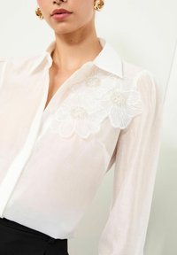 Camisa blanca de botones fabricada con un tejido ligero, que presenta tres flores bordadas con acentos de cuentas en el hombro izquierdo. Mangas largas.