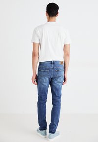Jeans de mezclilla azul con corte recto, que cuentan con bolsillos traseros y una etiqueta de piel de la marca. Combinados con una camiseta blanca sencilla y zapatos claros.
