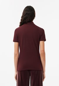 Polo shirt bordeaux in cotone, con colletto classico e maniche corte. Texture liscia con orlo arrotondato e dettagli minimi.