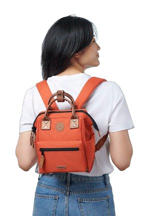 Woman with black hair in white shirt wearing an orange mini backpack with brown handles and black zippers, shown from the back.