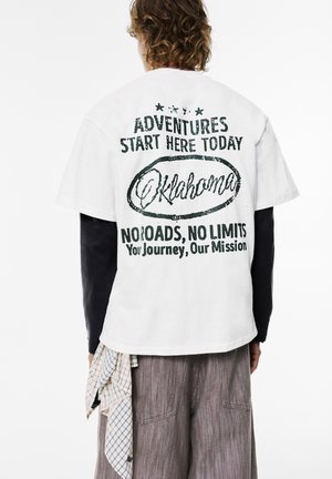 Hvid bomulds t-shirt med grøn trykt tekst: "Eventyr Starter Her I Dag Oklahoma" med et cirkulært design og yderligere sætninger.