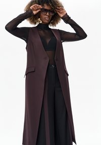 Długa, bezrękawnikowa kamizelka w bordowym kolorze z ostrymi klapami, nałożona na przezroczystą czarną bluzkę, zestawiona z czarnymi spodniami. Duże czarne okulary przeciwsłoneczne dopełniają stylizację.