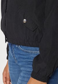 Svart lett jakke med elastiske mansjetter, med en sidelomme med en sølvknapp, parret med blå denimjeans.