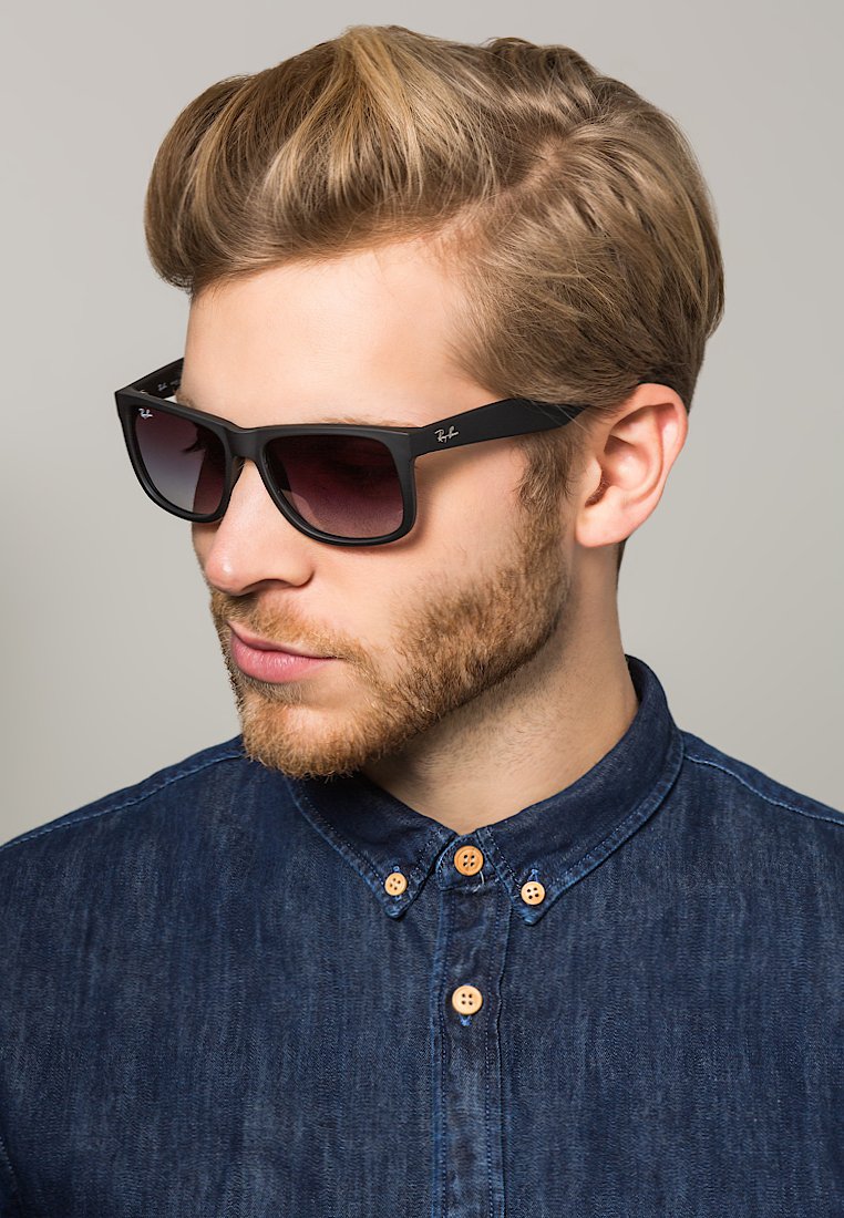 Occhiali da sole neri a forma rettangolare con lenti sfumate, con il marchio Ray-Ban sul telaio. Indossati con una camicia di jeans.