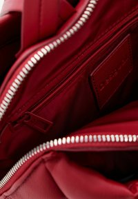 Interior de un bolso de piel roja con cremallera plateada, que presenta un logotipo en relieve y múltiples compartimentos para una mejor organización. Forro de tela texturizada.