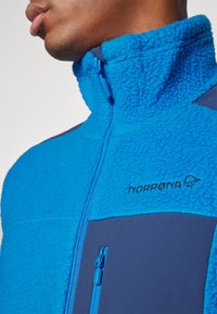 Blue fleece jacket with high collar, textured main body, contrasting navy panels, and embroidered logo. Features zippered pocket.