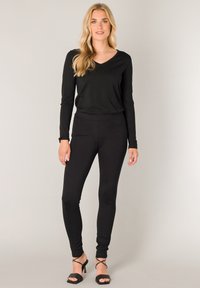 Zwarte lange mouwen V-hals top gecombineerd met zwarte strakke leggings. Beide hebben een zachte textuur. Het model draagt zwarte strapless hakken.
